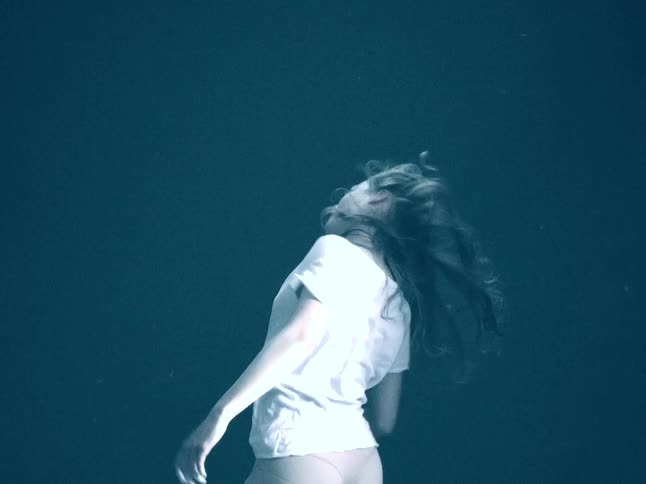 A woman wearing a white shirt and white powder on her forehead performs energetic and expressive movements as her long hair flows and flips against a solid dark background in a dim studio