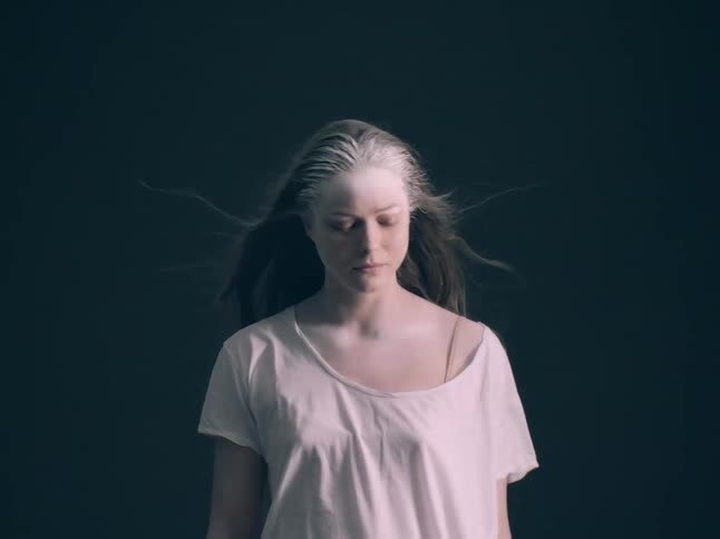 A woman wearing a loose white shirt with white powder covering her face and hair stands still as her long hair blows in the wind against a solid dark background in a dim studio
