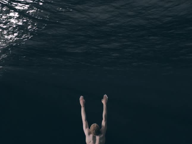 A view from behind shows a shirtless man with short blonde hair slowly raising both arms toward the dark surface of deep water from below