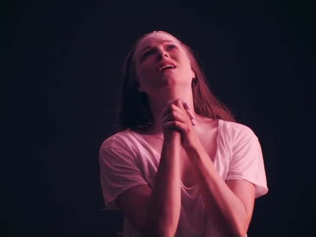 A young woman wearing a pink shirt stands indoors with her hands clasped together in sadness looking up as if praying