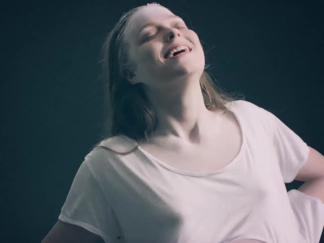 A woman wearing a white shirt with white powder on her face and hair tilts her head back and laughs against a solid dark background in a dim studio