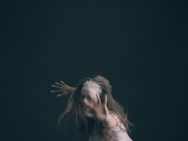 A woman with white paint on her forehead performs an intense expressive dance against a dark backdrop she moves her arms and hair with frantic energy reaching toward the camera with a strained facial expression