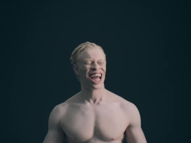 A shirtless man with blonde hair and pale skin laughs and shouts while looking toward the camera against a solid dark background in a studio setting