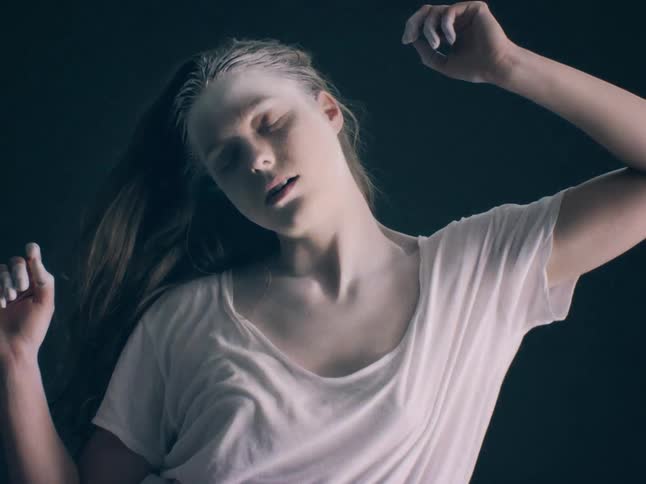 A young woman with white powder on her face and hair performs an expressive dance against a dark background