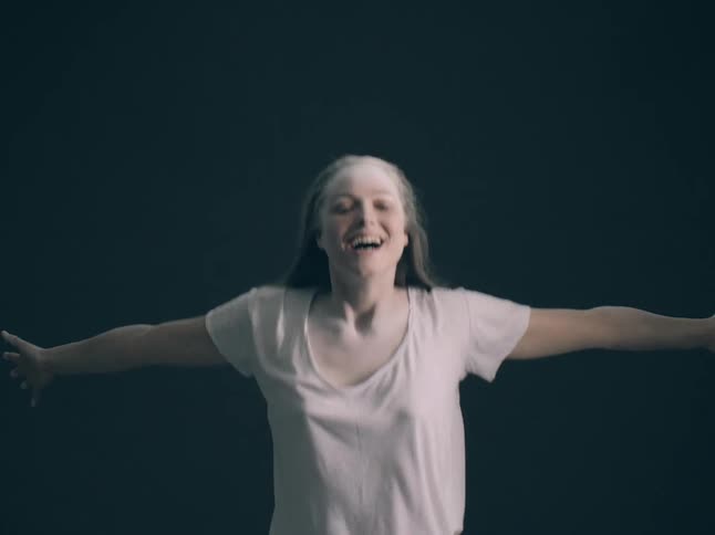 A young woman with powder on her face and hair jumps joyfully against a dark background she has her arms outstretched and a wide ecstatic smile as she moves through the air