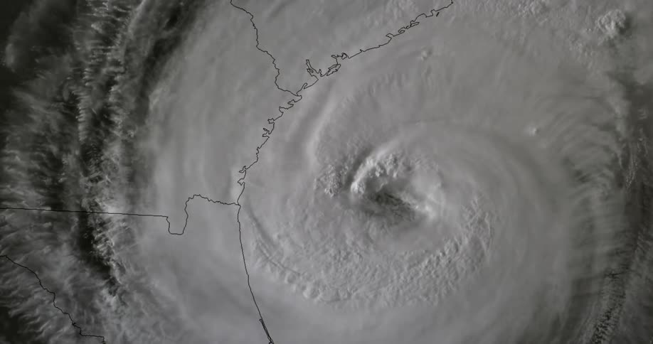 A black and white satellite view shows the large eye of a hurricane swirling near a coastline with map outlines visible beneath the thick cloud formations
