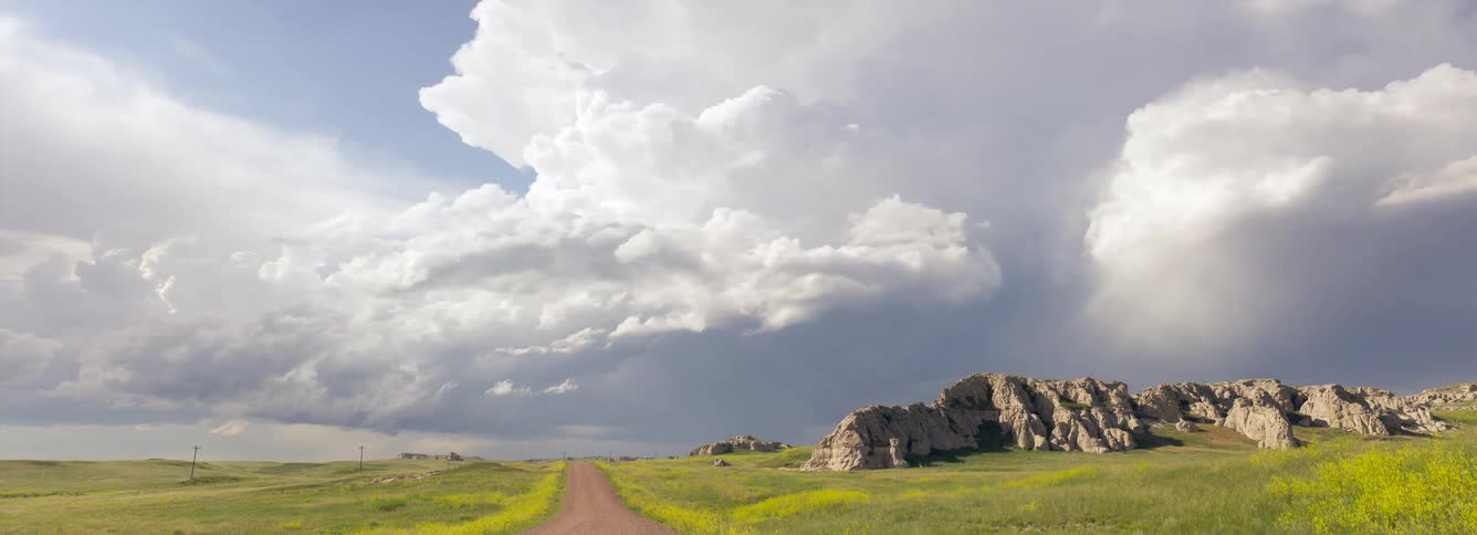 A view from a dirt road shows massive white and grey cumulus clouds building over green fields and rocky hills under a bright sky