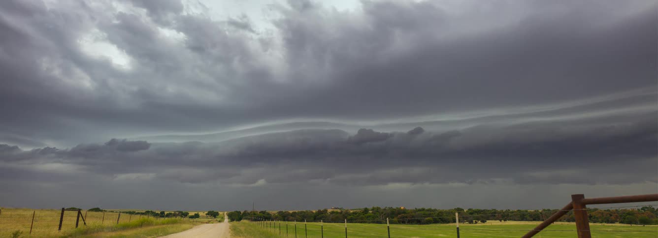 Large grey storm clouds with a rotating shelf formation move quickly over a rural dirt road and green fields under a dark and overcast sky