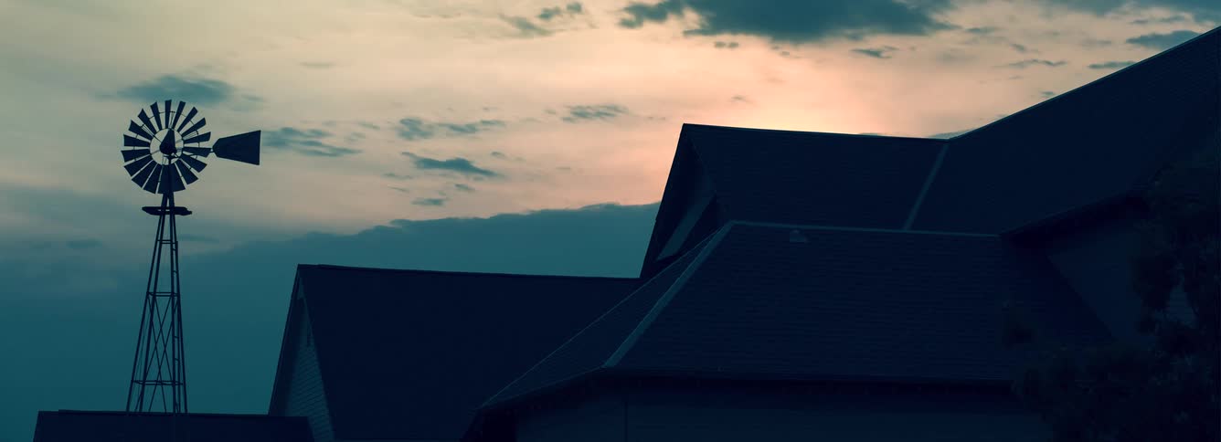 A metal windmill stands in silhouette next to the dark rooftops of suburban houses against a pale orange and teal sky with scattered clouds during a quiet sunset