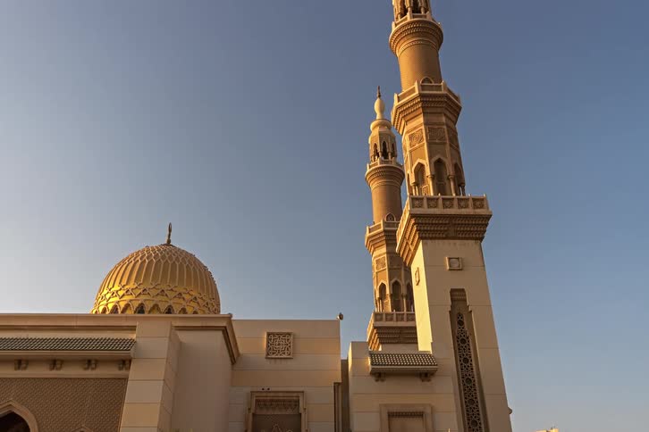 A low angle view shows a large mosque with a gold dome and two tall tan minarets against a clear blue sky during a bright sunset