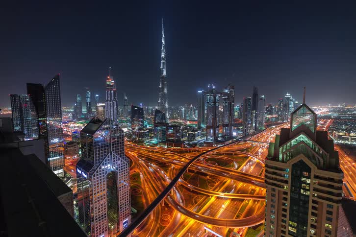 A high angle view at night shows the bright city lights of dubai with the burj khalifa towering over illuminated skyscrapers and a complex network of highways filled with glowing orange traffic trails