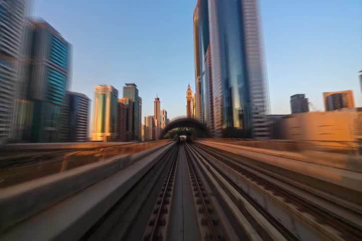 A point of view time lapse shows a train traveling on elevated tracks through a modern city with tall skyscrapers and glass buildings under a clear blue sky during a bright sunset