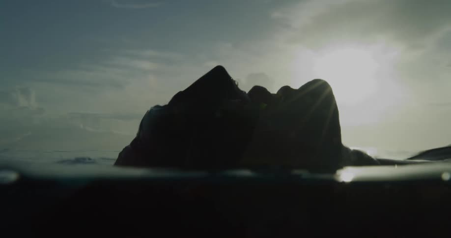 Person lying in shallow water with their face above the surface as waves ripple under a cloudy sky