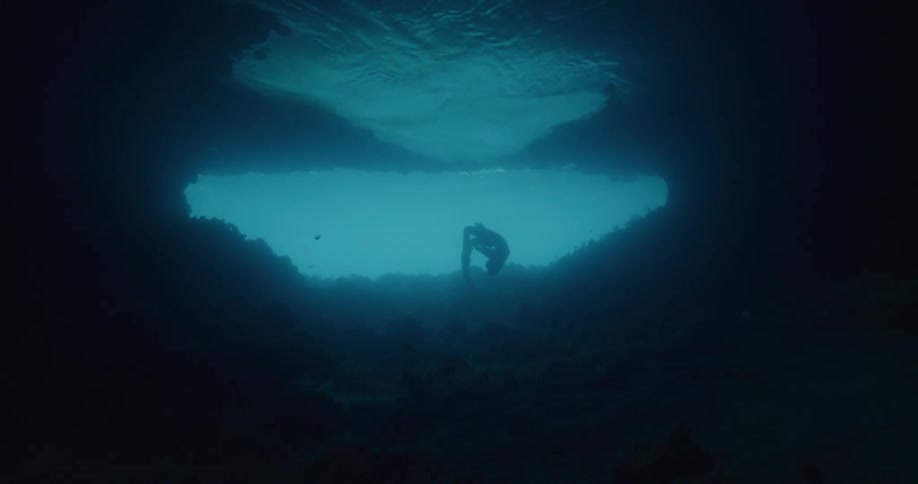 A silhouetted freediver gracefully swims through a dark underwater cavern toward a bright opening of blue light the diver performs a slow midwater somersault while moving through the deep shadows of the rocky cave