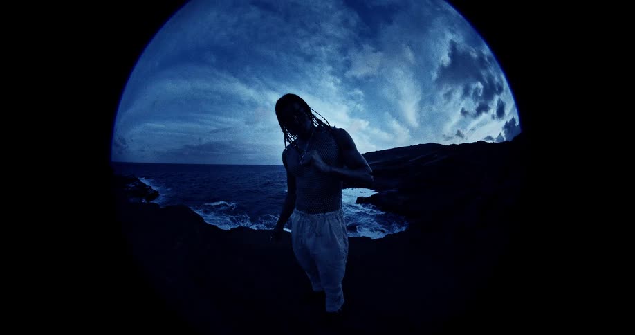 A person with long braided hair is shown through a circular lens as they stand on a rocky shore next to the ocean under a dark blue sky with bright clouds