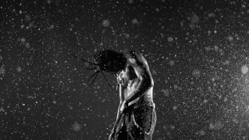 Bare chested man stands outdoors in the evening while it rains