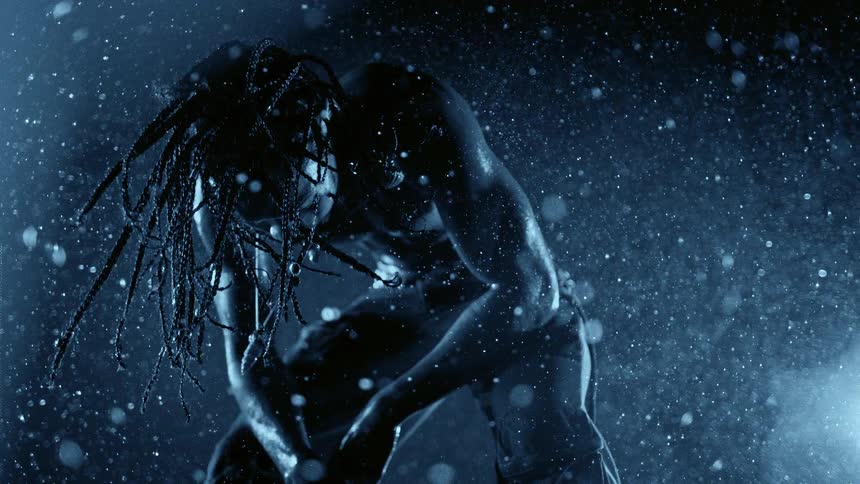 A person with long braided hair and jewelry on their face is seen in a blue light while water droplets fall and glisten against a dark background