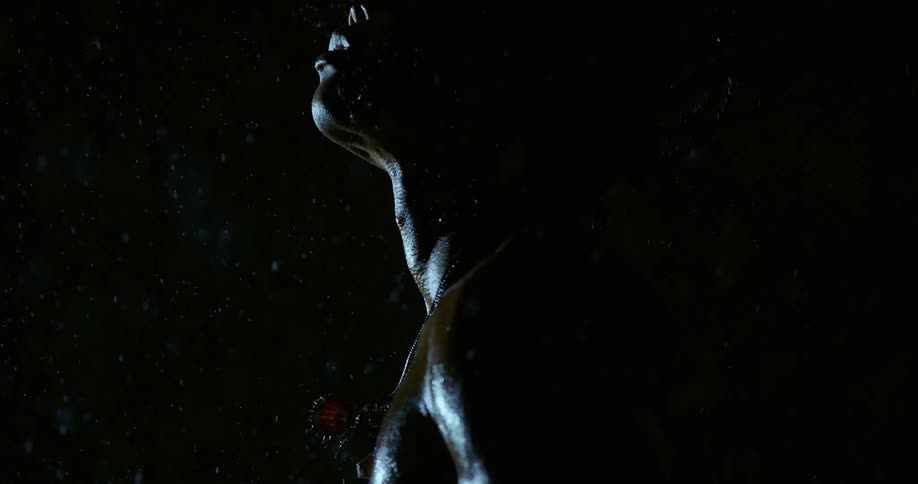 A person is shown in profile tilting their head back toward the sky as rain falls heavily around them the scene uses dark highcontrast lighting that highlights the water droplets glistening on their skin and shoulders