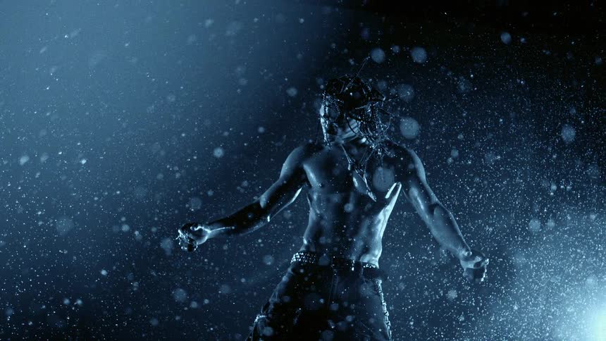 A person with long braided hair and jewelry on their face is seen in a blue light while water droplets fall and glisten against a dark background
