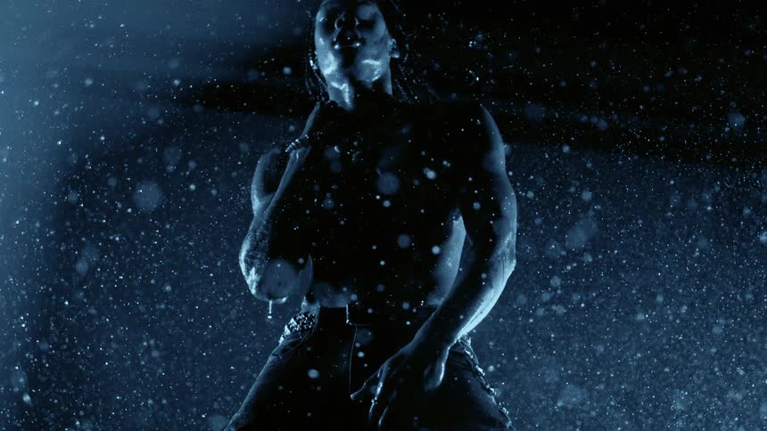 Bare chested man stands outdoors at night in stylish pants while it rains