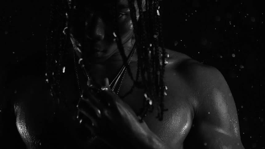 A man with braided hair and necklaces stands in a dark setting as water droplets fall onto his bare shoulders and chest while he moves his hand toward his neck in a close view
