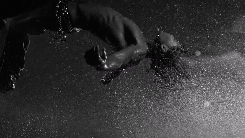 A shirtless man with long wet hair performs a backflip through a heavy spray of water against a black background