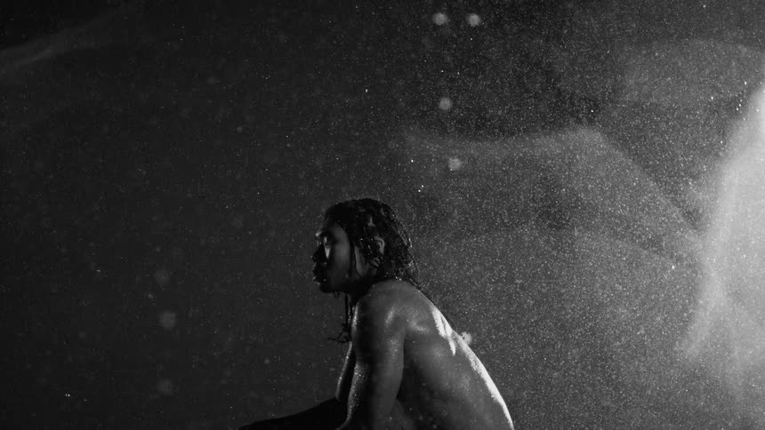 A muscular man with long hair sits in a dark space while a heavy spray of water falls over his back and shoulders in a backlit black and white scene where droplets glisten against the shadows