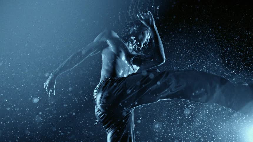 Shirtless man performs a jumping move under rain with blue lighting and visible water particles