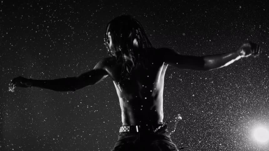 A shirtless young man with dreadlocks standing outdoors in the rain at night