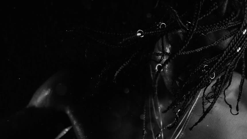 A person with long braided hair and facial jewelry is shown in a black and white close view while water droplets fall and glisten against a dark background