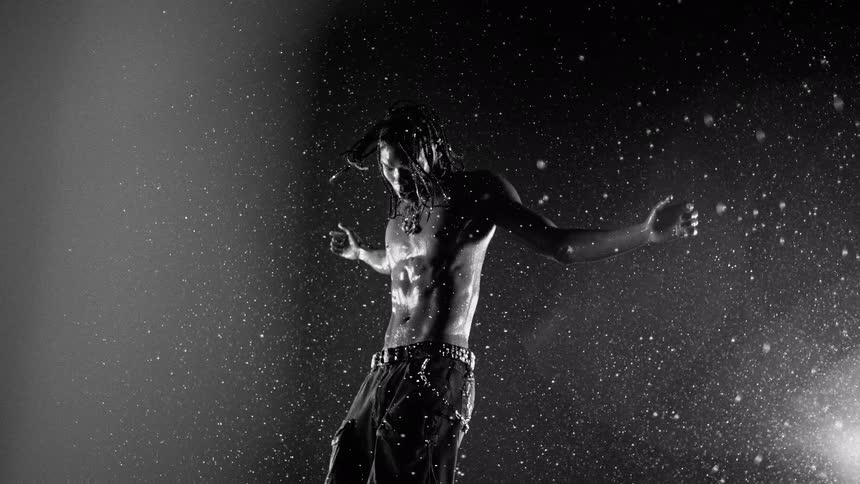 A shirtless man with braided hair stands with his arms outstretched as water droplets fall around him in dramatic blackandwhite lighting