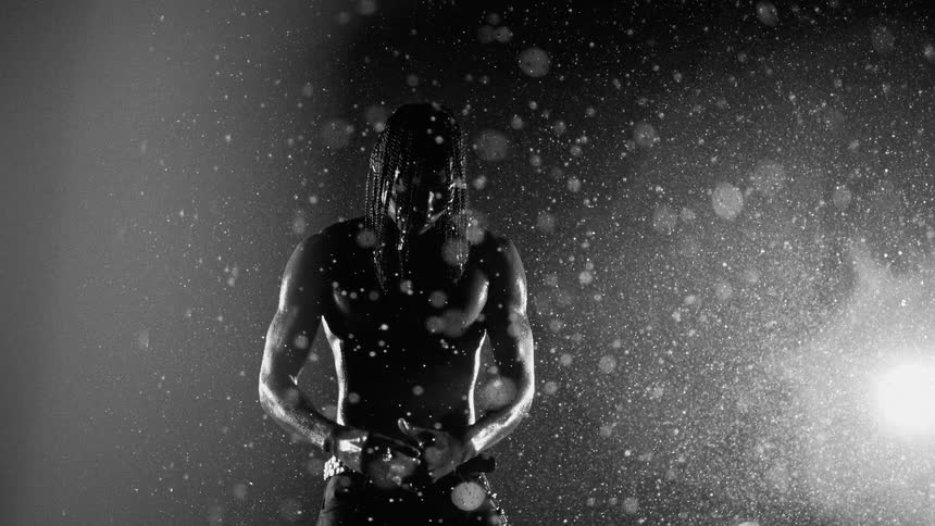 A shirtless man with long braids stands in a dark studio under falling water