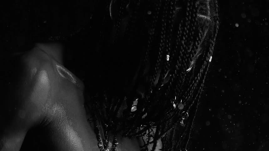 In this blackandwhite video a person with long braids stands amidst falling water droplets that glisten against the dark backgroundthe person is shown from a low angle highlighting their wet skin and the various metallic rings or charms woven into
