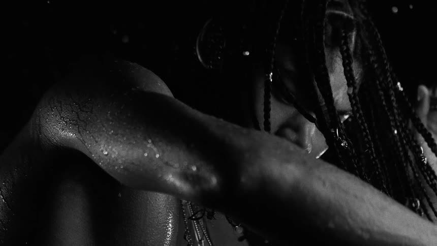 The video shows a closeup blackandwhite shot of a person with long braided hair covered in water droplets they move their arm across their face in a slow dramatic motion while rain or water falls against a dark background
