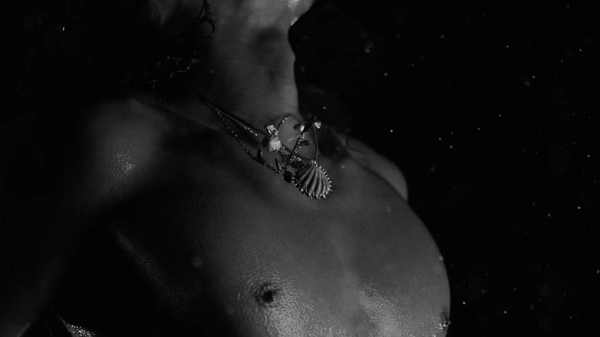 A dramatic blackandwhite closeup shows a persons neck and collarbone adorned with layered necklaces illuminated against a dark background with floating particles