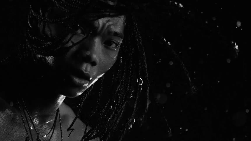 Man with braids under heavy rain with face partially illuminated against a dark background