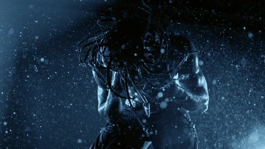 Light shines on a bare chested muscular man turning his head as the braids in his hair move and raindrops fill the air