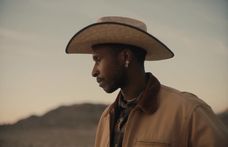 Man wearing a cowboy hat stands on rural land with mountains in the distance