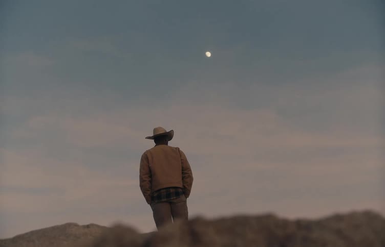 Person wearing a hat looks at the sky at dusk with the moon visible above