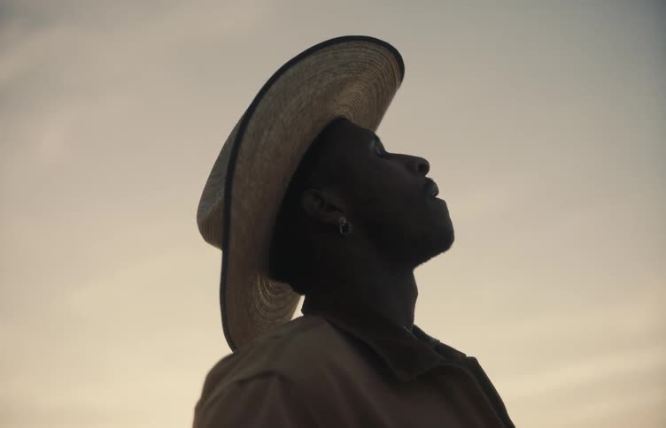 A man in a straw cowboy hat stands in silhouette against a soft golden sunset skyhe tilts his head back with his eyes closed appearing to take a deep reflective breath