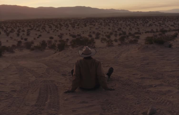 Man wearing a hat sitting on the sand observes a desert landscape at dusk with mountains in the background