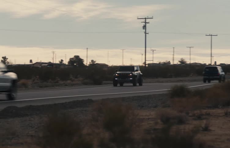 A black suv drives along a winding desert highway during a cloudy evening while utility poles and sparse vegetation line the road under a pale sky