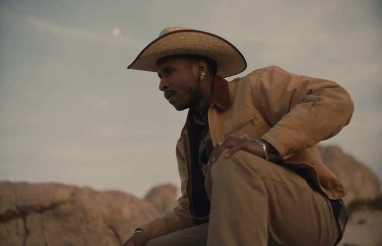 Person wearing a hat crouching in an arid landscape at dusk with the moon visible in the sky