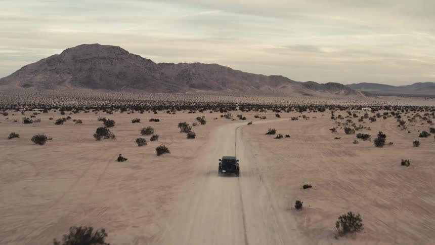 An aerial shot follows a dark offroad vehicle as it travels down a straight dirt path through a vast arid desert plain scattered shrubs dot the sandy landscape which leads toward a large rugged mountain range under a pale evening sky