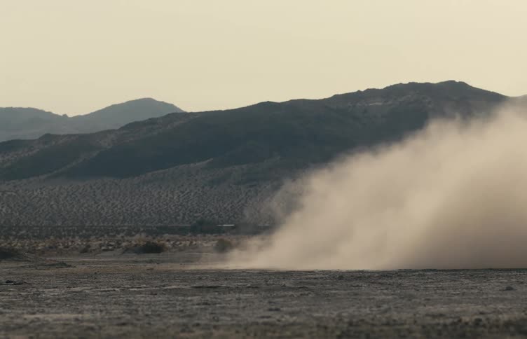 A large cloud of dust rises across a barren desert plain with mountains in the background