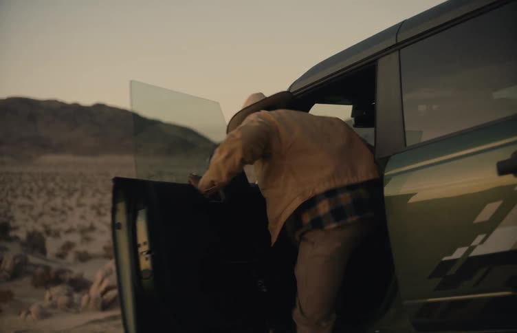A man wearing a brown jacket and a cowboy hat opens the door of an suv parked in the wilderness and steps inside at sunset