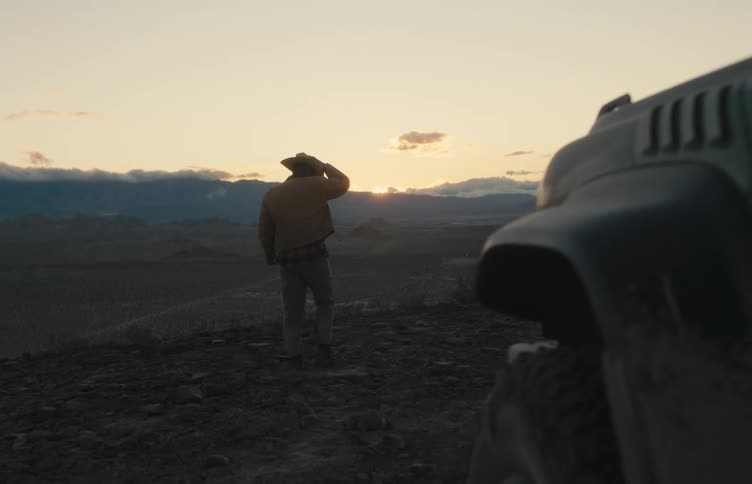 The video captures a lone man in a cowboy hat standing on a rocky overlook at sunset next to a parked offroad vehicle he gazes out across a vast mountainous desert landscape as the light fades behind the horizon