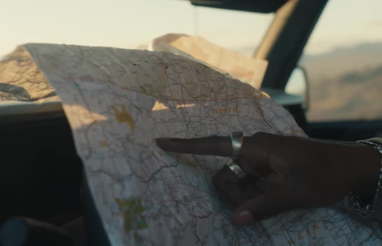 A person wearing silver rings on their fingers holds and points at a paper map while sitting inside a vehicle during a bright sunset in the desert
