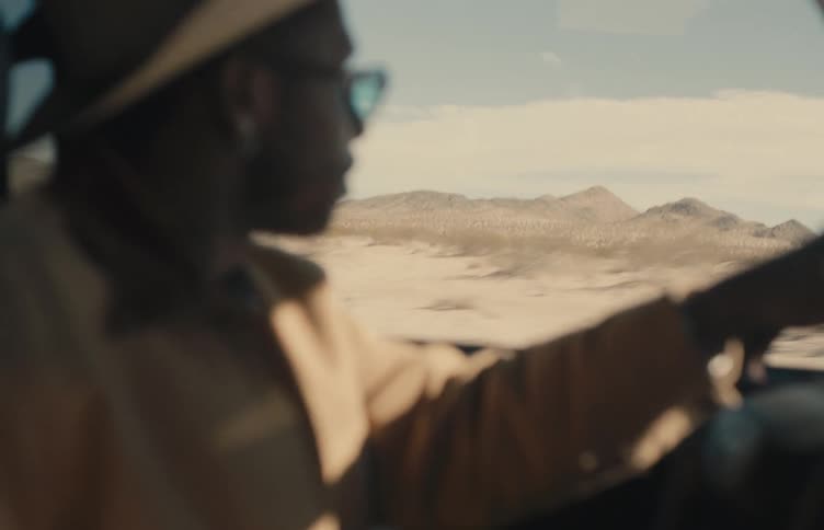 A blurred profile of a black man wearing a hat and sunglasses driving a car through a desert landscape with mountains in the distance