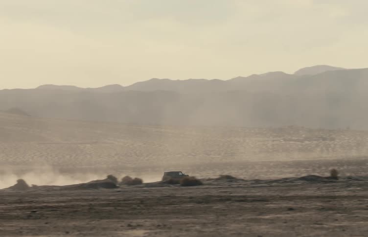 A distant view shows a dark vehicle driving across a flat desert landscape and kicking up a trail of dust against a backdrop of hazy mountains and a pale sky